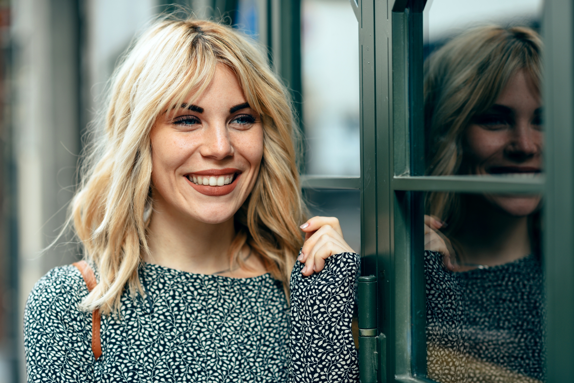 A woman with shoulder-length blonde hair smiles while standing by a window with a reflection of her visible in the glass. She is wearing a black and white patterned top and holds the edge of the window frame, exuding a cheerful and relaxed demeanor.