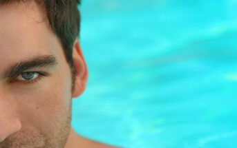 Close-up of the left half of a man's face with stubble, set against a blurred turquoise background, which appears to be water. The man's green eye is prominently visible, with an intense and focused gaze. The rest of his face is partially out of the frame.
