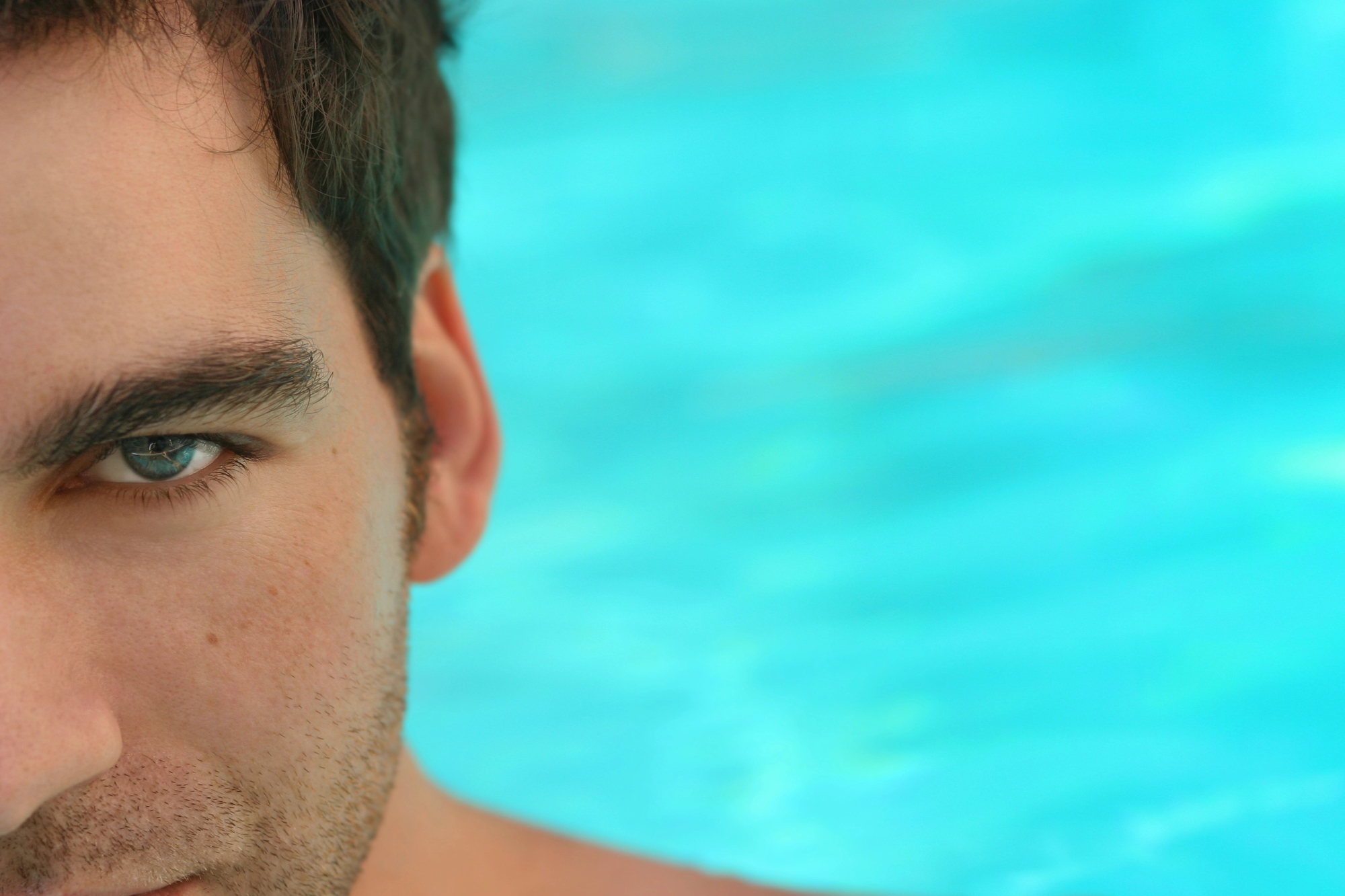 Close-up of the left half of a man's face with stubble, set against a blurred turquoise background, which appears to be water. The man's green eye is prominently visible, with an intense and focused gaze. The rest of his face is partially out of the frame.