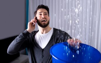 A man wearing a cardigan over a white shirt holds a phone to his ear with one hand while holding a blue bucket with the other hand. Water is pouring from the ceiling into the bucket. His facial expression appears stressed. Vertical blinds are in the background.
