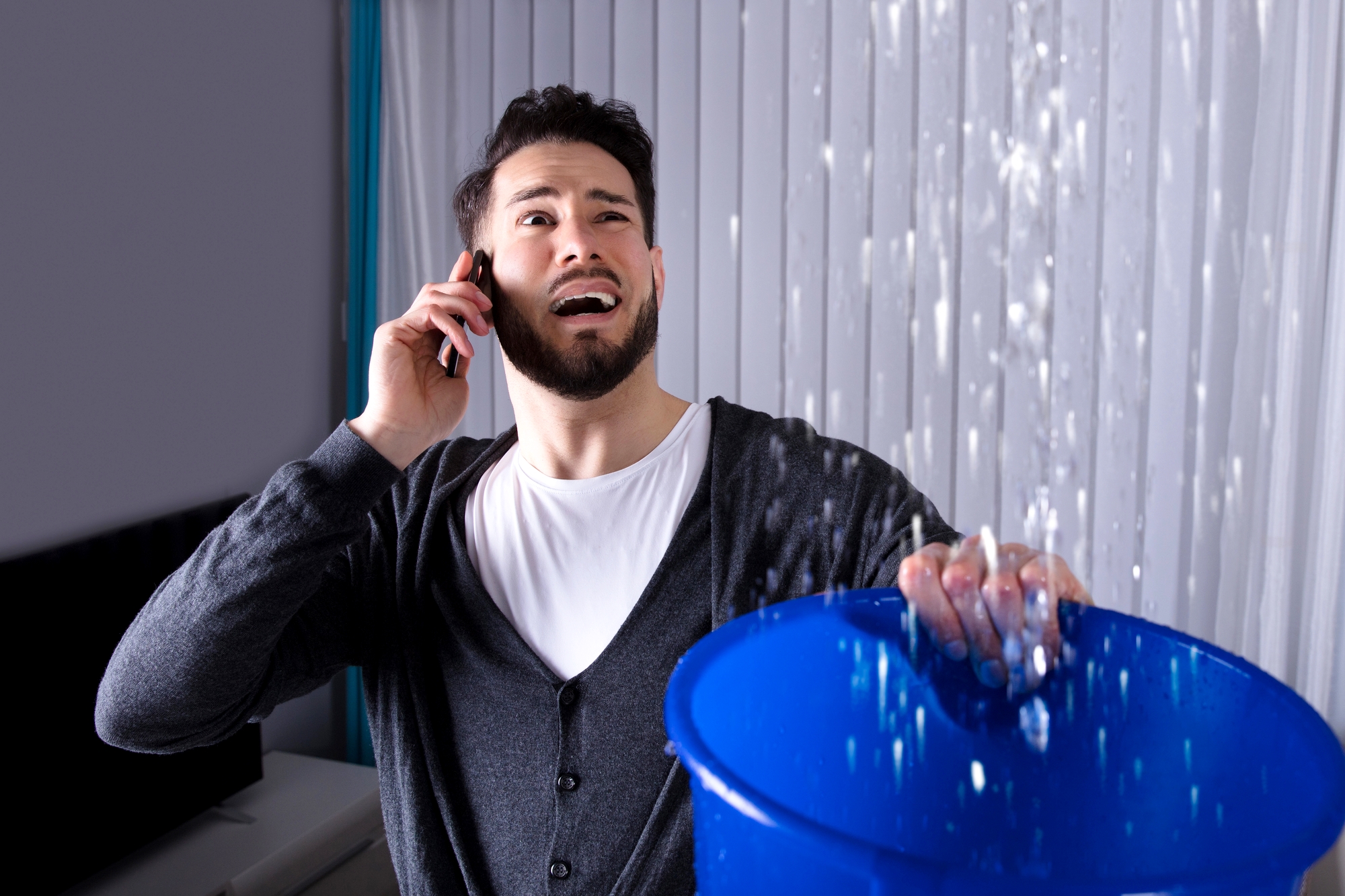 A man wearing a cardigan over a white shirt holds a phone to his ear with one hand while holding a blue bucket with the other hand. Water is pouring from the ceiling into the bucket. His facial expression appears stressed. Vertical blinds are in the background.