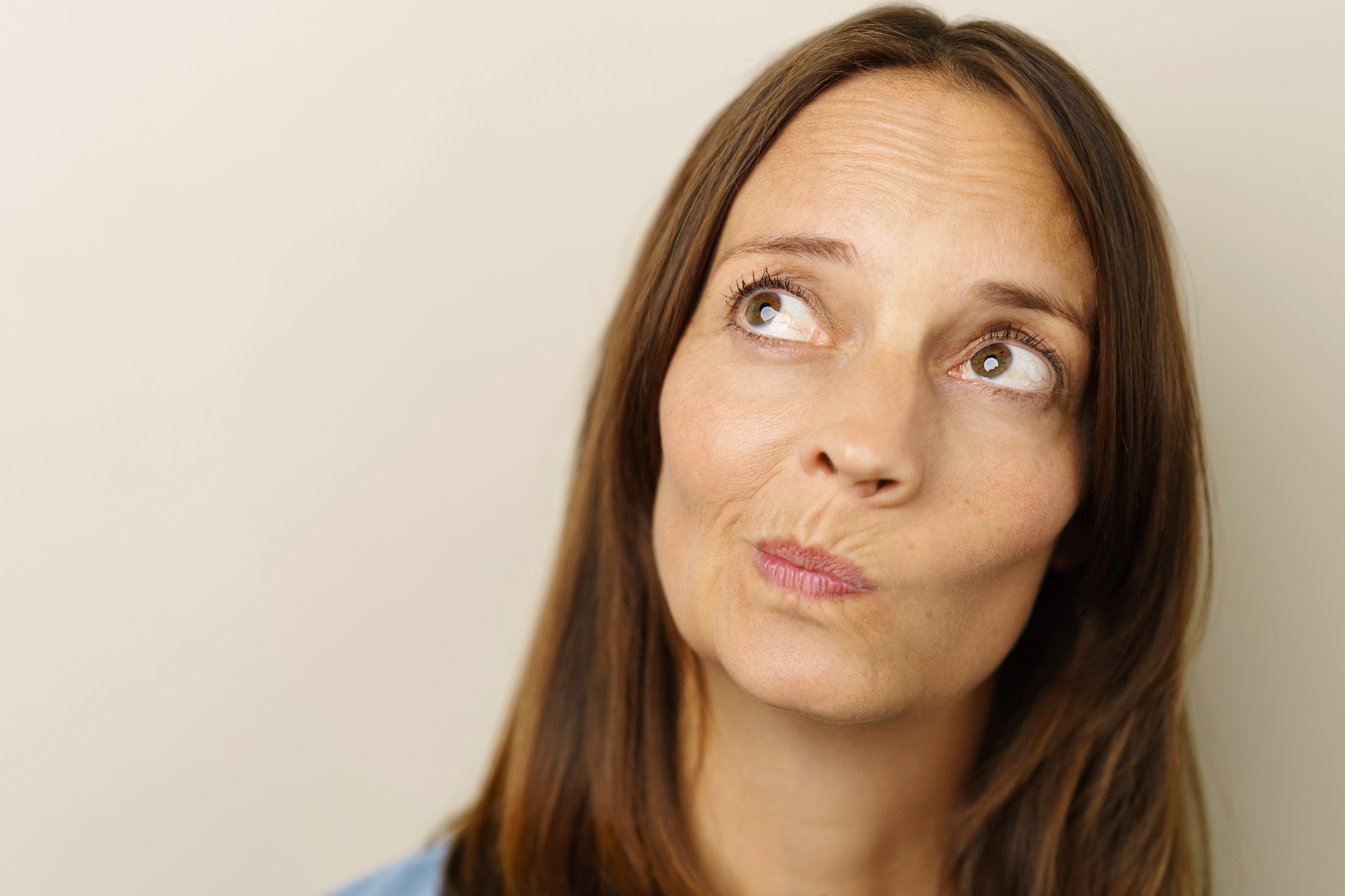 A woman with long brown hair is looking up and to the side with a thoughtful expression on her face. She is wearing a light blue top, and the background is a plain, light-colored wall.