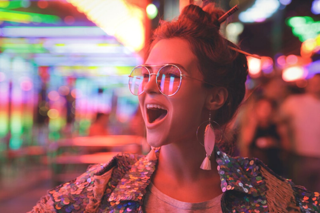 A person with a joyful expression stands in a vibrant, neon-lit environment. They are wearing reflective aviator sunglasses, feather earrings, and a sequined jacket, with their hair stylishly tied up. The background is filled with colorful, blurry lights.