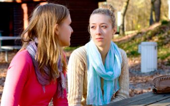 Two young women are sitting at a wooden picnic table outside in a wooded area. One woman with long hair and a pink top is talking, while the other with blonde hair and a blue scarf is listening attentively. Sunlight filters through the trees, casting a warm glow.