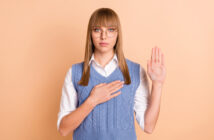 A person with straight blonde hair, wearing glasses, a white shirt, and a blue knit vest, stands against a peach-colored background. They have one hand raised and the other placed over their chest, appearing to take an oath or pledge.