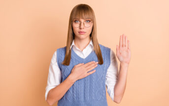 A person with straight blonde hair, wearing glasses, a white shirt, and a blue knit vest, stands against a peach-colored background. They have one hand raised and the other placed over their chest, appearing to take an oath or pledge.