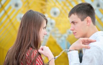 A man and a woman stand facing each other in front of a bright yellow geometric background, both with serious expressions. Their wrists are cuffed together with handcuffs, binding them closely. The man wears a white shirt, and the woman wears a red patterned top.