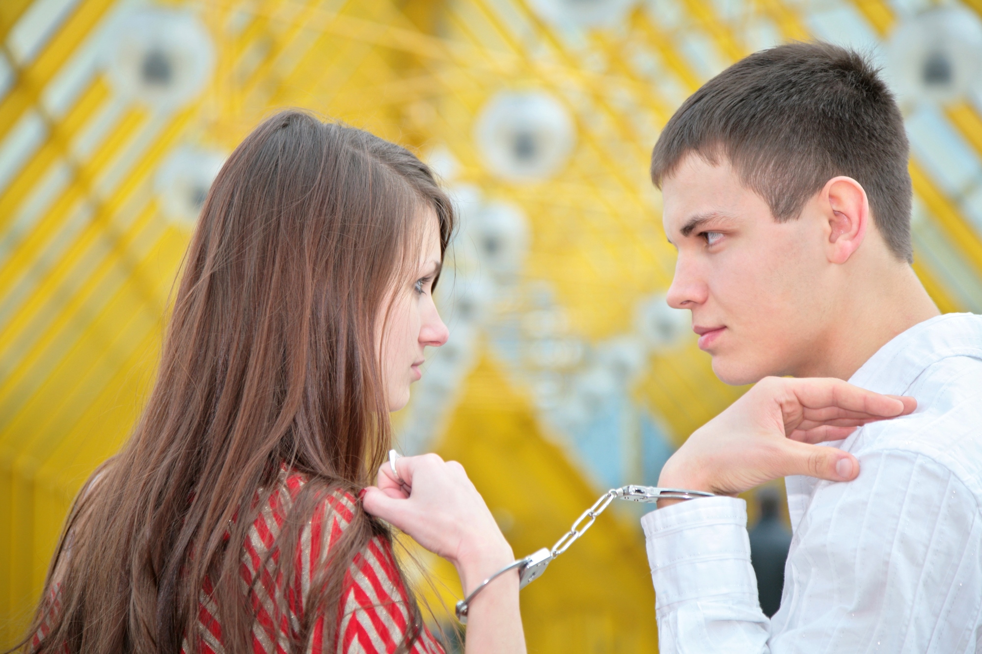 A man and a woman stand facing each other in front of a bright yellow geometric background, both with serious expressions. Their wrists are cuffed together with handcuffs, binding them closely. The man wears a white shirt, and the woman wears a red patterned top.