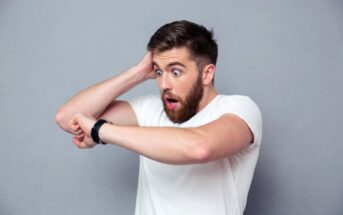 A man with a beard and short hair wearing a white t-shirt looks surprised while checking the time on his wristwatch. He has one hand on his head and a shocked expression on his face, standing against a plain grey background.