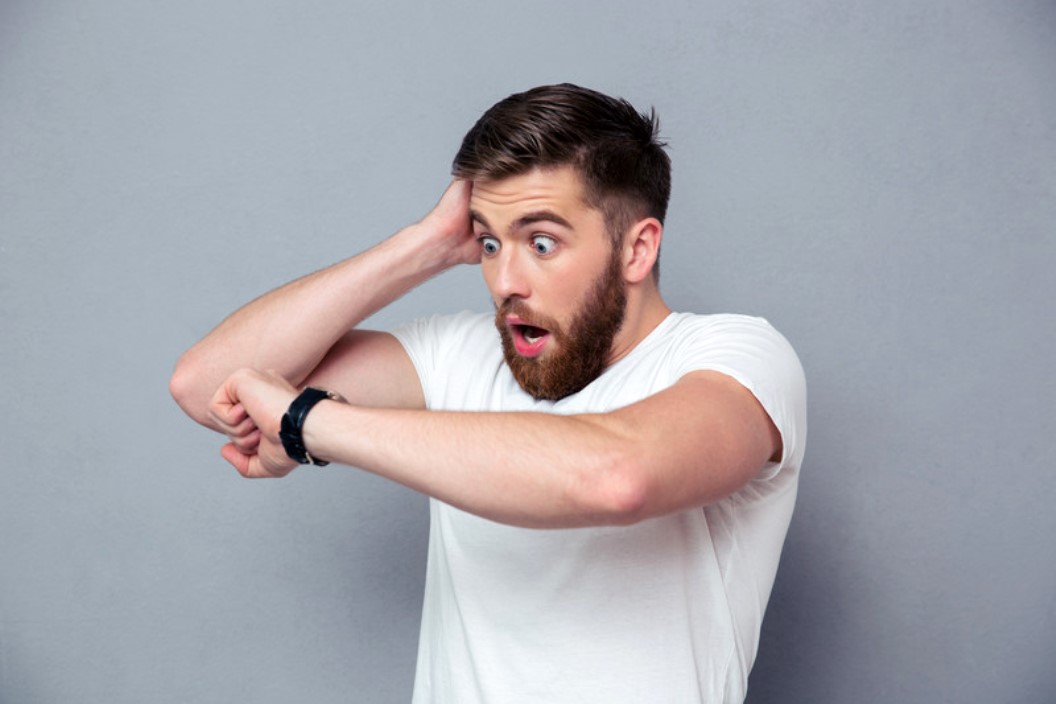 A man with a beard and short hair wearing a white t-shirt looks surprised while checking the time on his wristwatch. He has one hand on his head and a shocked expression on his face, standing against a plain grey background.