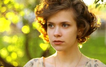 A young person with short, curly hair looks off into the distance with a thoughtful expression. The background features a softly blurred, sunlit scene with vibrant green foliage and circular light bokeh.