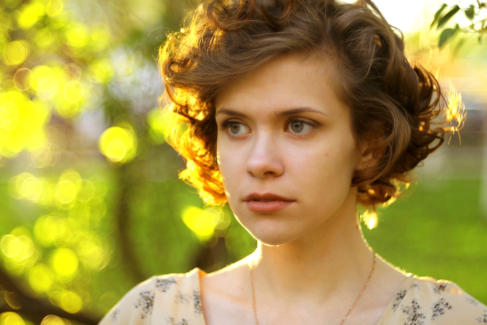 A young person with short, curly hair looks off into the distance with a thoughtful expression. The background features a softly blurred, sunlit scene with vibrant green foliage and circular light bokeh.