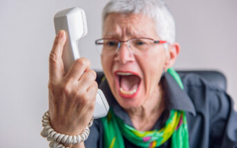 An elderly person with short gray hair, wearing glasses and a colorful scarf, is angrily holding a landline telephone receiver and shouting into it. The background is blurred, focusing attention on the person's expressive face and gesture.