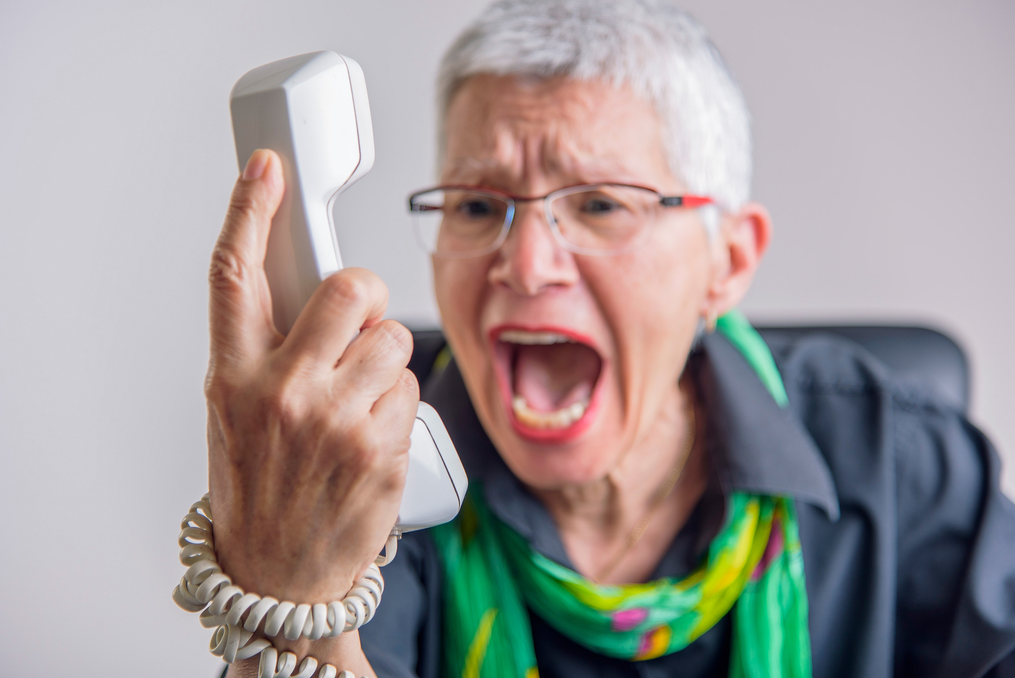 An elderly person with short gray hair, wearing glasses and a colorful scarf, is angrily holding a landline telephone receiver and shouting into it. The background is blurred, focusing attention on the person's expressive face and gesture.