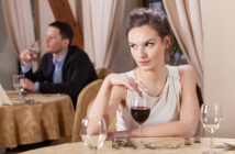 A woman in a white dress sits at a table with a glass of red wine, looking to the side with a disinterested expression. In the background, a man in a suit is seated at another table, holding a wine glass and appearing thoughtful. Both are in a dimly lit restaurant.