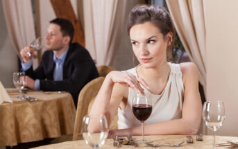 A woman in a white dress sits at a table with a glass of red wine, looking to the side with a disinterested expression. In the background, a man in a suit is seated at another table, holding a wine glass and appearing thoughtful. Both are in a dimly lit restaurant.