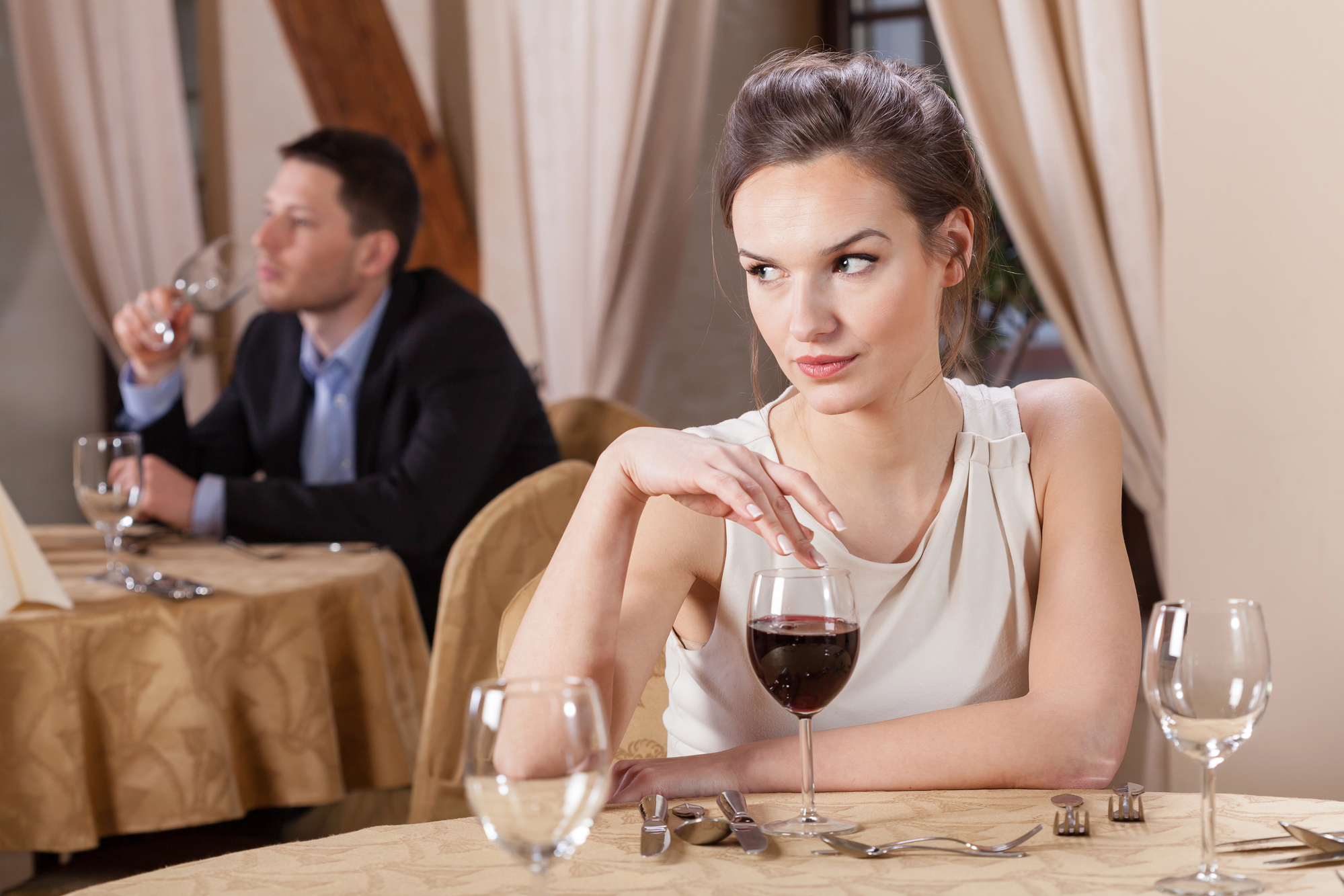 A woman in a white dress sits at a table with a glass of red wine, looking to the side with a disinterested expression. In the background, a man in a suit is seated at another table, holding a wine glass and appearing thoughtful. Both are in a dimly lit restaurant.