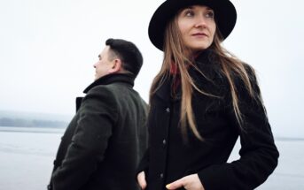 Two people standing outdoors near a body of water on a cloudy day. The person in the foreground, wearing a black coat and a black hat, looks off into the distance. The person in the background, also in a dark coat, faces away, adding a sense of solitude.