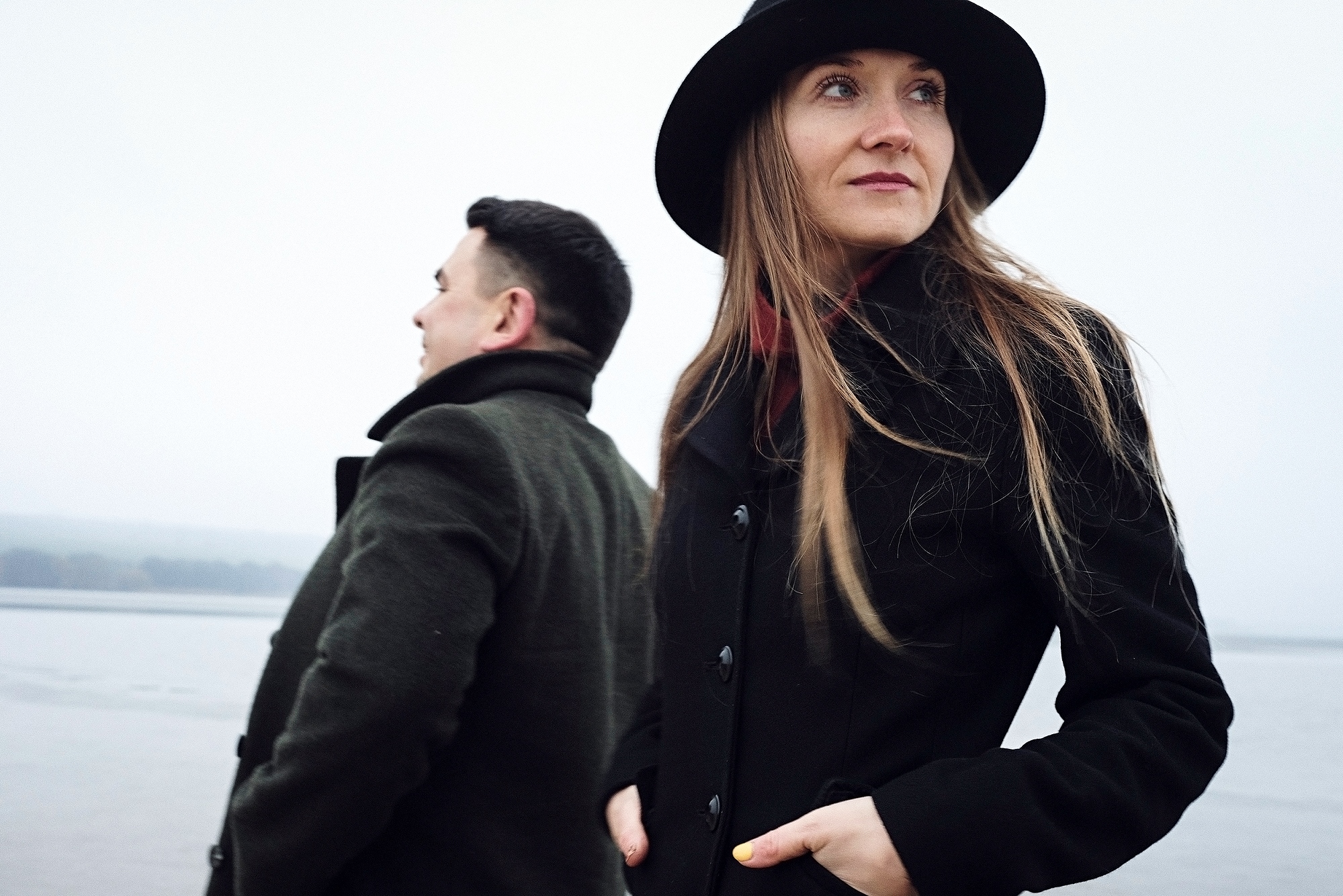 Two people standing outdoors near a body of water on a cloudy day. The person in the foreground, wearing a black coat and a black hat, looks off into the distance. The person in the background, also in a dark coat, faces away, adding a sense of solitude.