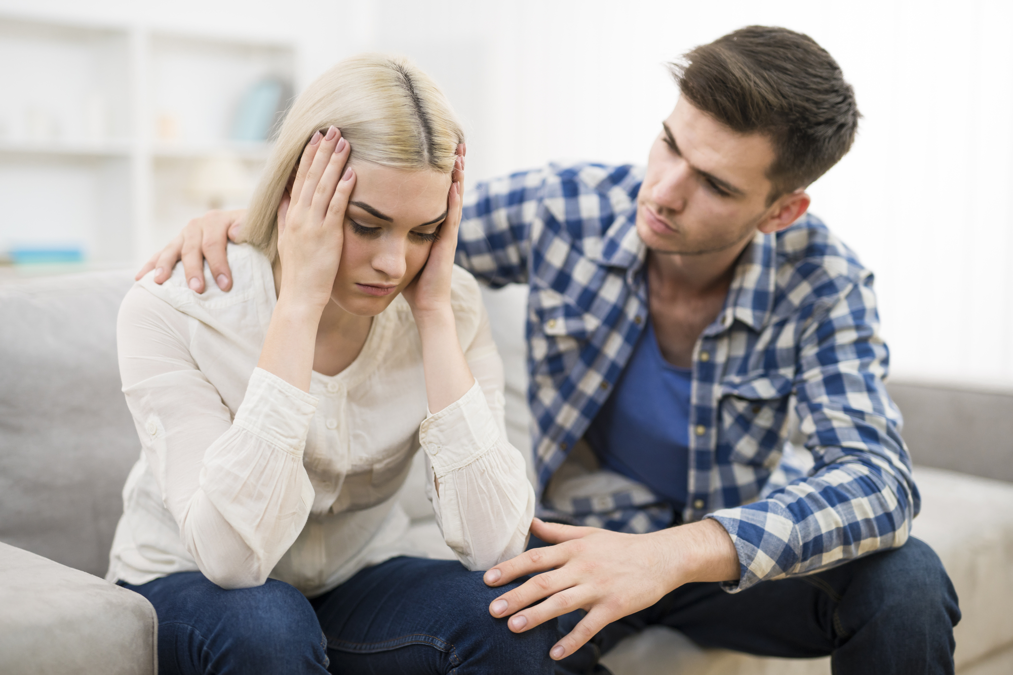 A man in a blue plaid shirt sits beside a distressed woman with blond hair on a couch. He has his arm around her and his hand on her knee, offering comfort as she holds her head in her hands. They appear to be having a serious conversation.