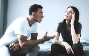 A couple sits on a bed in an argument. The man on the left, wearing a white t-shirt, gestures emphatically with one hand. The woman on the right, wearing a green t-shirt, appears distressed, resting her head in one hand while looking upwards.