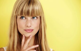 A woman with long blonde hair and bangs stands in front of a bright yellow background. She is looking slightly upward with a thoughtful expression, resting her chin on her hand. She is wearing a light-colored sleeveless top.