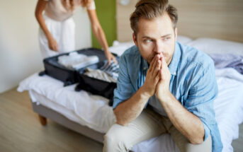 A man sits on the edge of a bed with his hands clasped near his mouth, looking pensive. In the background, a woman with curly hair is packing a suitcase on the bed. The room has light wooden flooring and a modern, minimalist design.