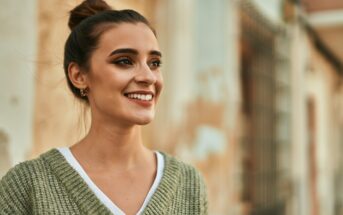 A young woman with dark hair tied in a bun smiles while looking to the side. She is wearing a green knit sweater over a white V-neck shirt. The background is blurry with warm-toned and pastel-colored walls.