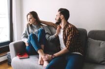 A woman and a man sit on a gray couch by a window. The woman, wearing a gray sweater and blue jeans, looks away, appearing unhappy. The man, in a plaid shirt and jeans, leans towards her, his arm around her shoulders, and attempts to comfort her. A red notebook and smartphone are on the couch beside them.