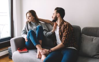 A woman and a man sit on a gray couch by a window. The woman, wearing a gray sweater and blue jeans, looks away, appearing unhappy. The man, in a plaid shirt and jeans, leans towards her, his arm around her shoulders, and attempts to comfort her. A red notebook and smartphone are on the couch beside them.