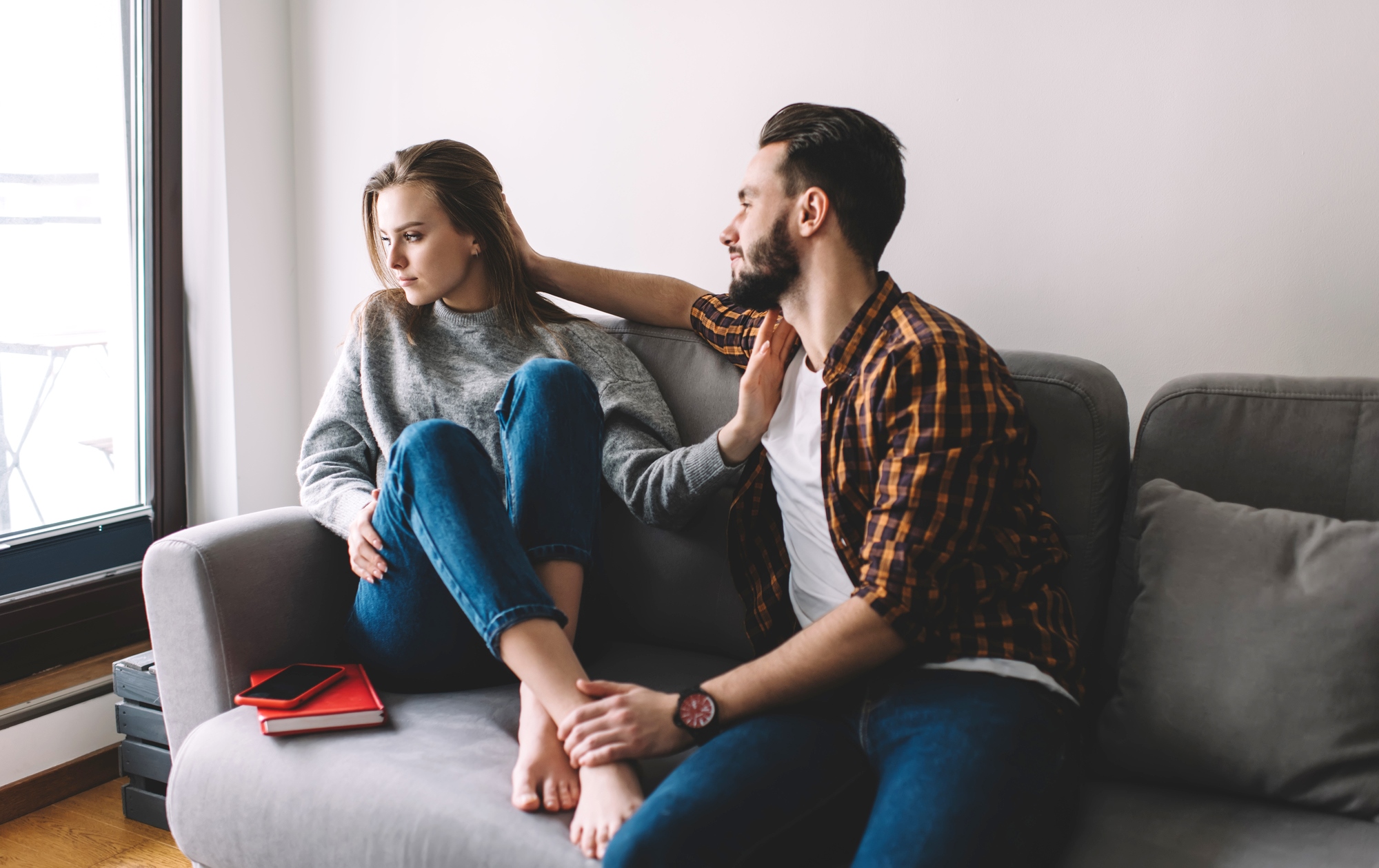 A woman and a man sit on a gray couch by a window. The woman, wearing a gray sweater and blue jeans, looks away, appearing unhappy. The man, in a plaid shirt and jeans, leans towards her, his arm around her shoulders, and attempts to comfort her. A red notebook and smartphone are on the couch beside them.