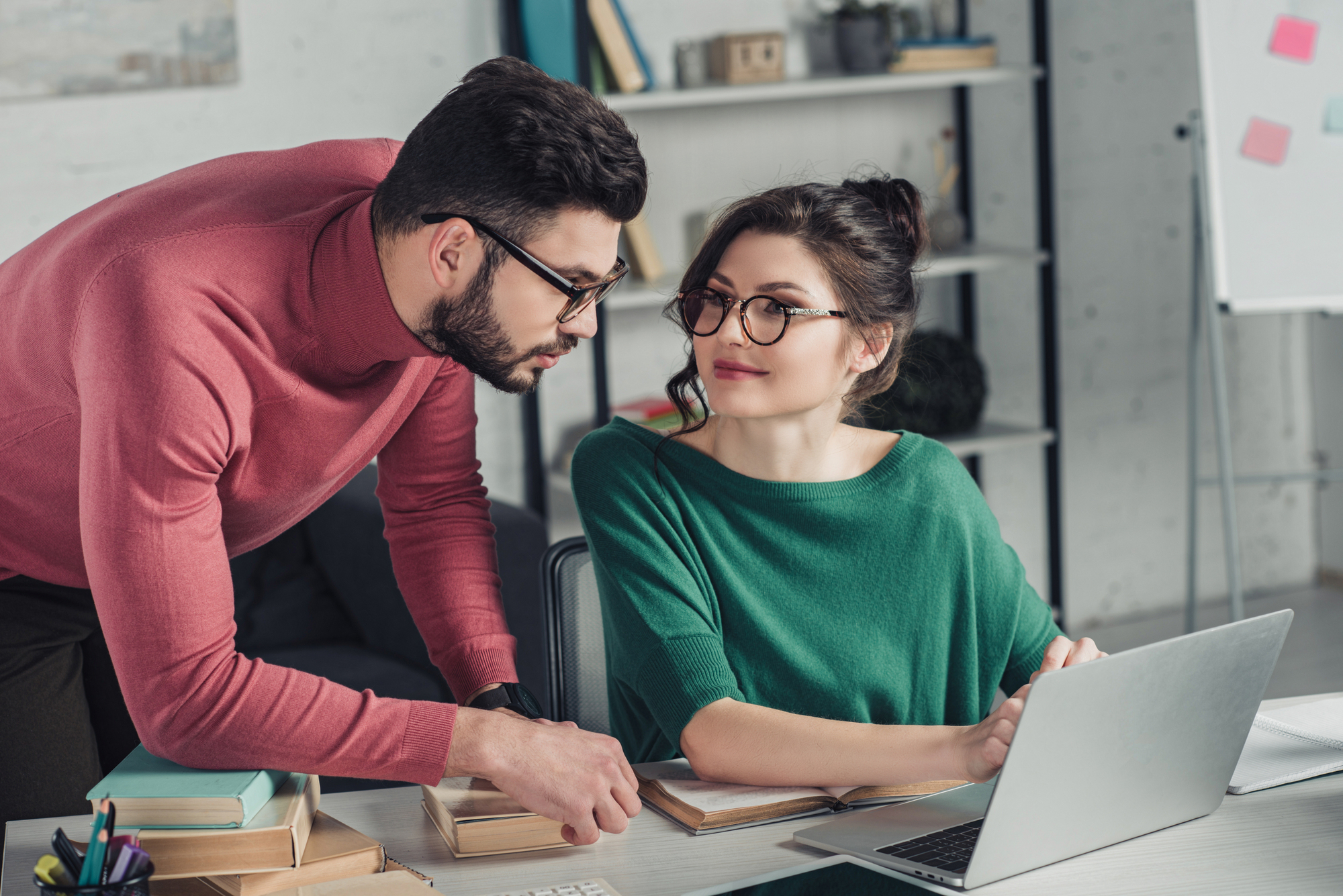 A man in a pink sweater leans over a desk to speak to a woman in a green sweater working on a laptop. Both are wearing glasses and appear to be in a modern office setting with shelves and books in the background. The woman looks up at the man attentively.