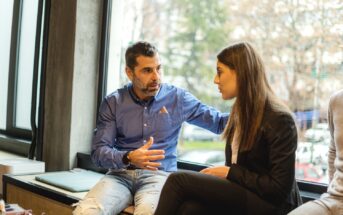 A man wearing a blue shirt and jeans is sitting next to a woman in business attire by a window. He appears to be in conversation with the woman, gesturing with one hand and resting his other hand on her shoulder. The background shows a blurred outdoor scene.