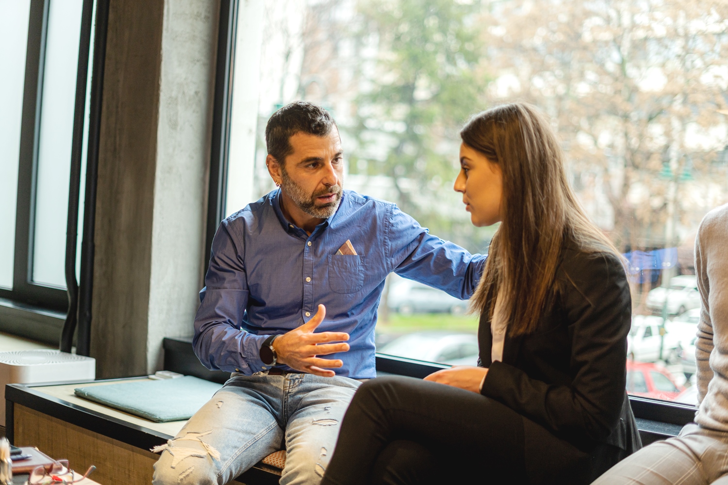 A man wearing a blue shirt and jeans is sitting next to a woman in business attire by a window. He appears to be in conversation with the woman, gesturing with one hand and resting his other hand on her shoulder. The background shows a blurred outdoor scene.