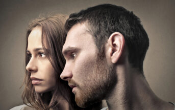 Close-up of a woman and a man standing side by side, both looking intently to the left. The woman has long brown hair and a serious expression, while the man has short brown hair, a beard, and a pensive look. The background is neutral, with soft lighting.