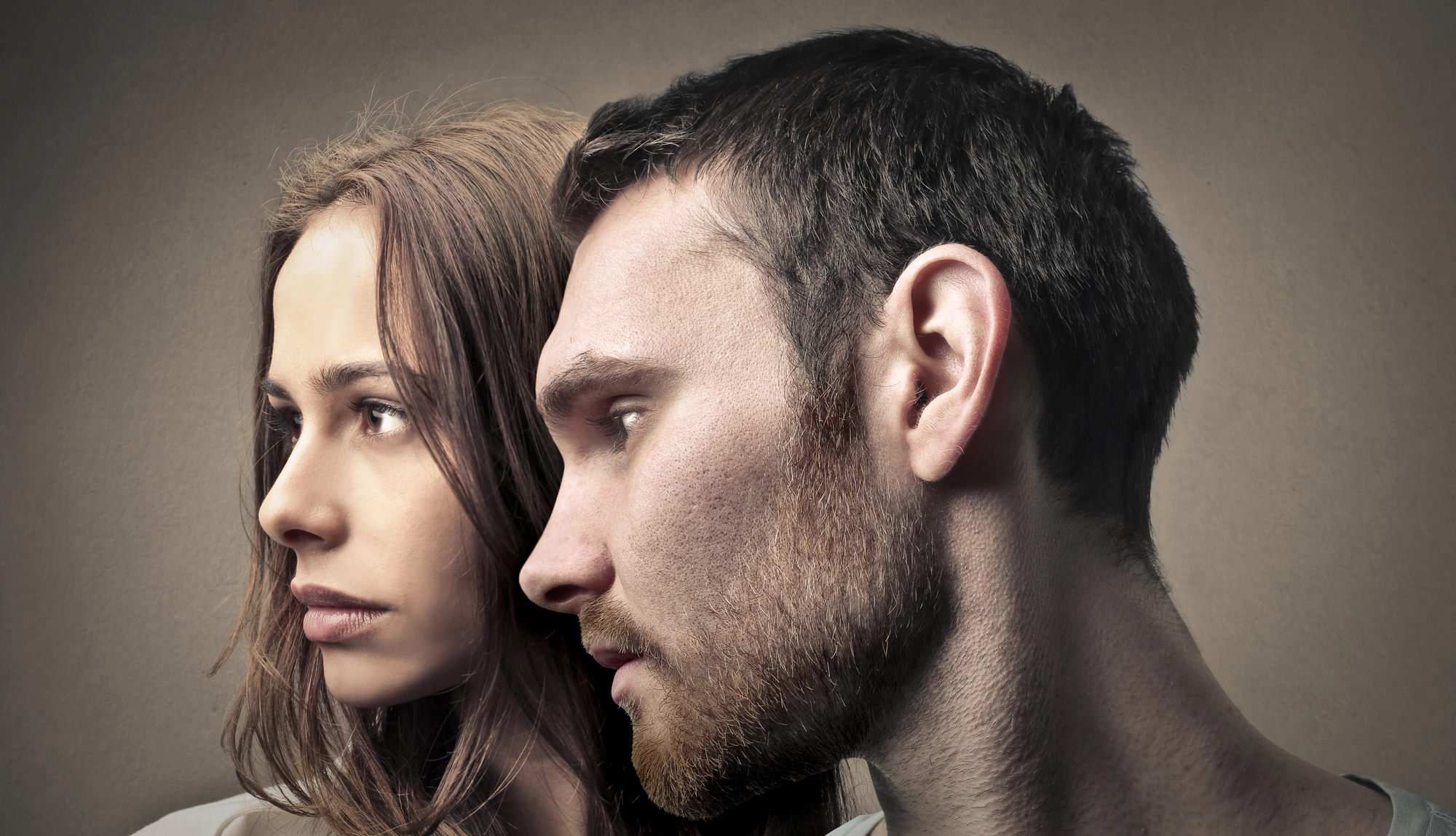 Close-up of a woman and a man standing side by side, both looking intently to the left. The woman has long brown hair and a serious expression, while the man has short brown hair, a beard, and a pensive look. The background is neutral, with soft lighting.