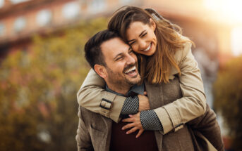 A smiling woman hugs a laughing man from behind as they enjoy a sunny day outdoors. They are both wearing coats, with autumn foliage and a building blurred in the background, creating a warm and cheerful atmosphere.