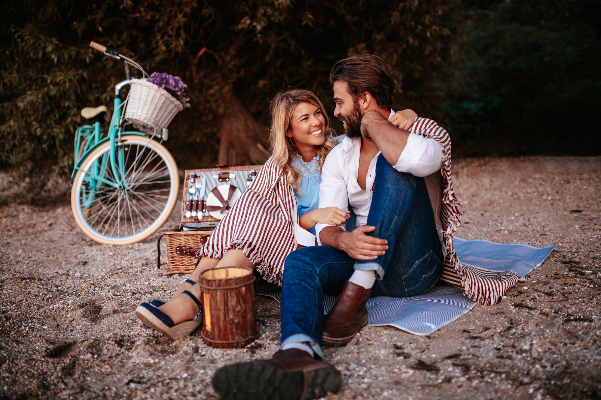 A couple sits on a beach blanket, smiling at each other. A bicycle with a flower-filled basket is parked nearby. They are surrounded by picnic items including a basket and small wooden bucket. The scene is relaxed and intimate with trees in the background.