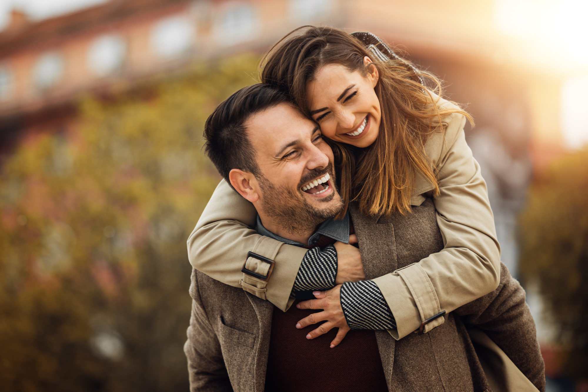 A smiling woman hugs a laughing man from behind as they enjoy a sunny day outdoors. They are both wearing coats, with autumn foliage and a building blurred in the background, creating a warm and cheerful atmosphere.