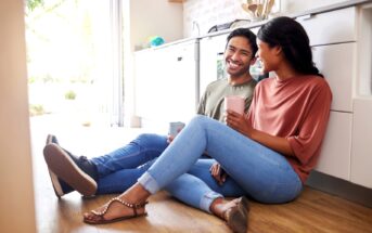 A joyful couple sits on a kitchen floor, smiling and enjoying a casual moment together with coffee mugs. The man, wearing a green shirt and jeans, sits next to the woman in a salmon top and jeans. Daylight streams in, highlighting the cozy, modern kitchen setting.