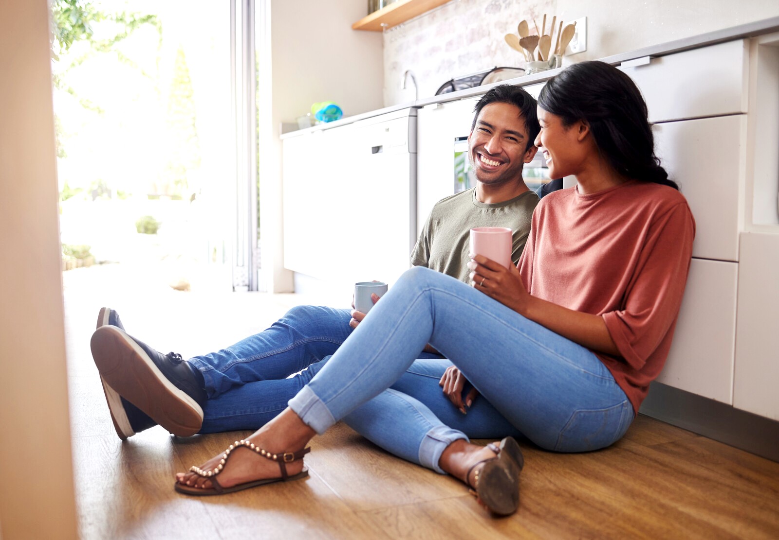 A joyful couple sits on a kitchen floor, smiling and enjoying a casual moment together with coffee mugs. The man, wearing a green shirt and jeans, sits next to the woman in a salmon top and jeans. Daylight streams in, highlighting the cozy, modern kitchen setting.