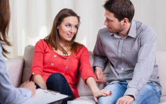A couple sits closely on a couch holding hands, engaging in a conversation. The woman wears a red sweater and the man a striped shirt. A person with a clipboard sits nearby, perhaps a therapist, indicating a counseling or therapy session.