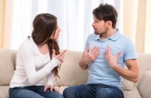 A woman and a man sitting on a couch are engaged in a heated discussion. The woman is pointing her finger, while the man has his hands placed on his chest, both with serious expressions. They are indoors with light curtains in the background.