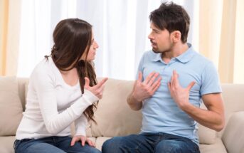 A woman and a man sitting on a couch are engaged in a heated discussion. The woman is pointing her finger, while the man has his hands placed on his chest, both with serious expressions. They are indoors with light curtains in the background.