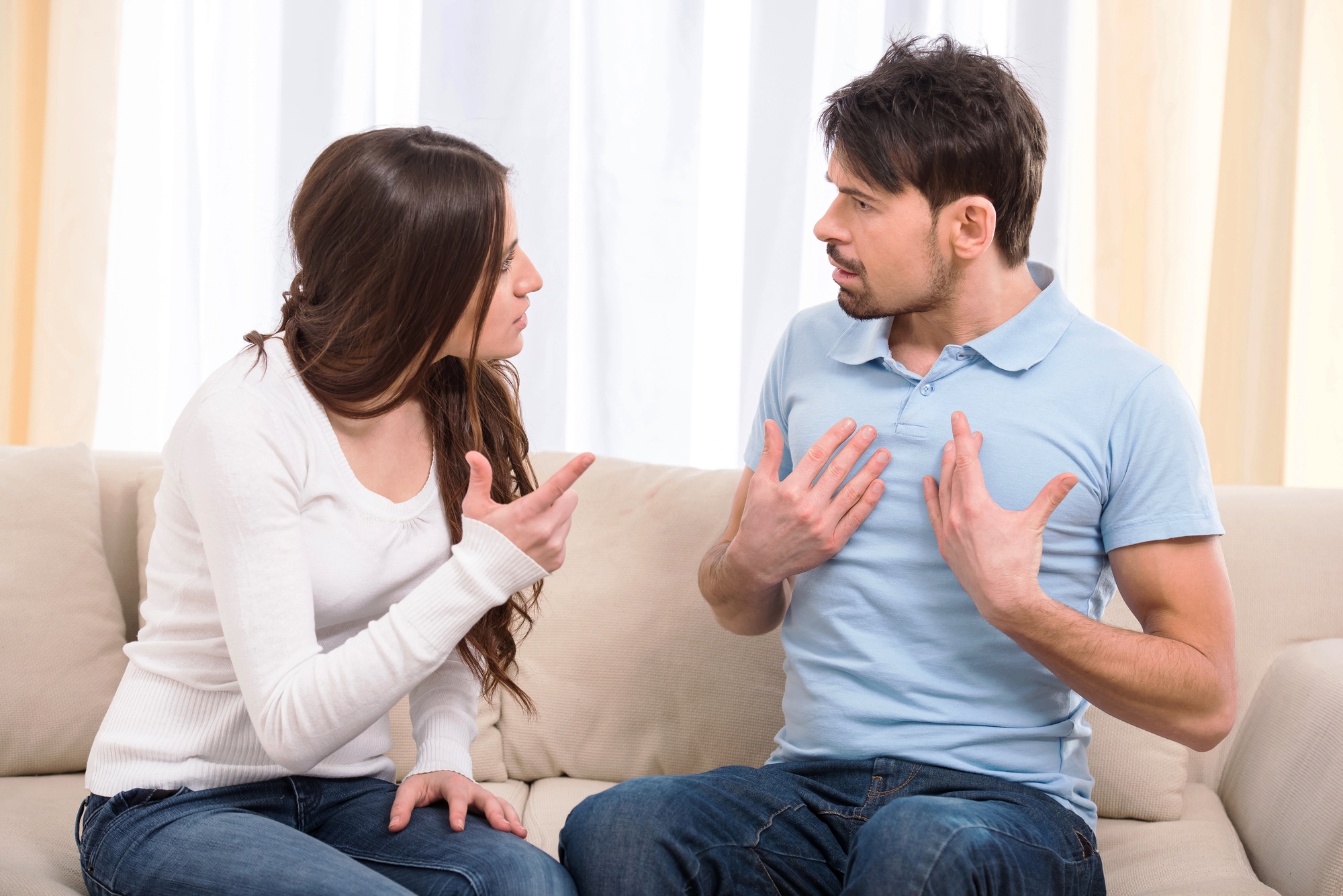 A woman and a man sitting on a couch are engaged in a heated discussion. The woman is pointing her finger, while the man has his hands placed on his chest, both with serious expressions. They are indoors with light curtains in the background.