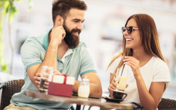A man with a beard and a woman wearing sunglasses are sitting at an outdoor café, smiling and enjoying drinks. The man is wearing a light blue polo shirt, and the woman is wearing a white T-shirt. They appear to be having a pleasant conversation.