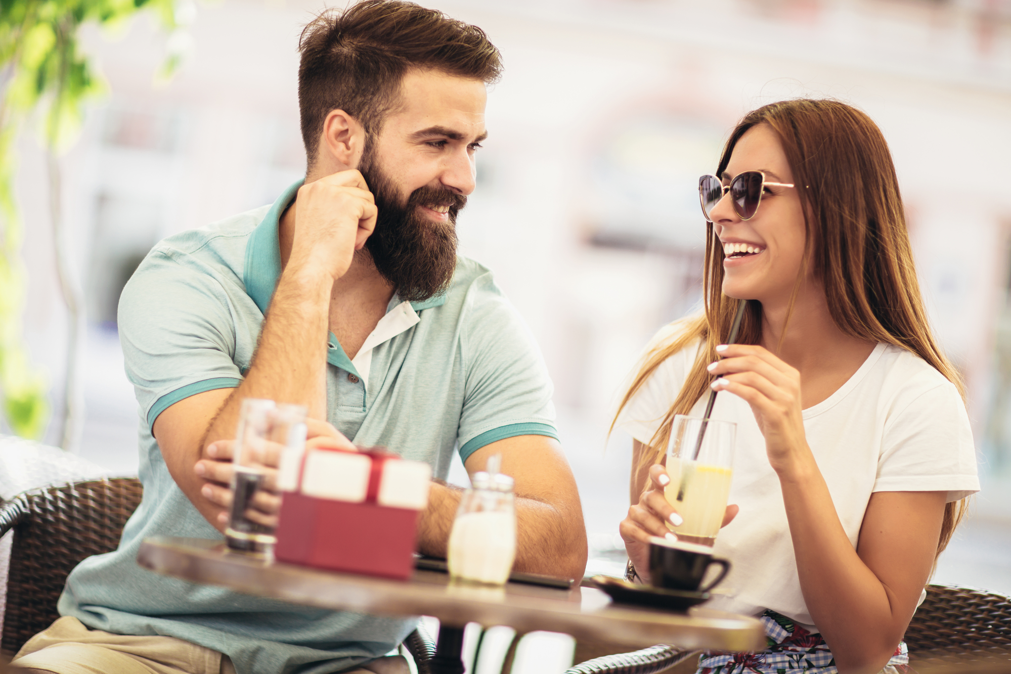 A man with a beard and a woman wearing sunglasses are sitting at an outdoor café, smiling and enjoying drinks. The man is wearing a light blue polo shirt, and the woman is wearing a white T-shirt. They appear to be having a pleasant conversation.