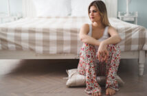 A woman sits on a pillow on the floor in front of a bed with a checkered bedspread. She is wearing a white tank top and floral pajama pants, with her arms resting on her knees and looking thoughtfully to the side. The room has a calm and serene atmosphere.
