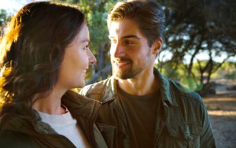 A man with short brown hair and a beard smiles at a woman with long brown hair in an outdoor setting. Both are dressed casually in green jackets, standing close together with trees and sunlight filtering through the background, creating a serene atmosphere.