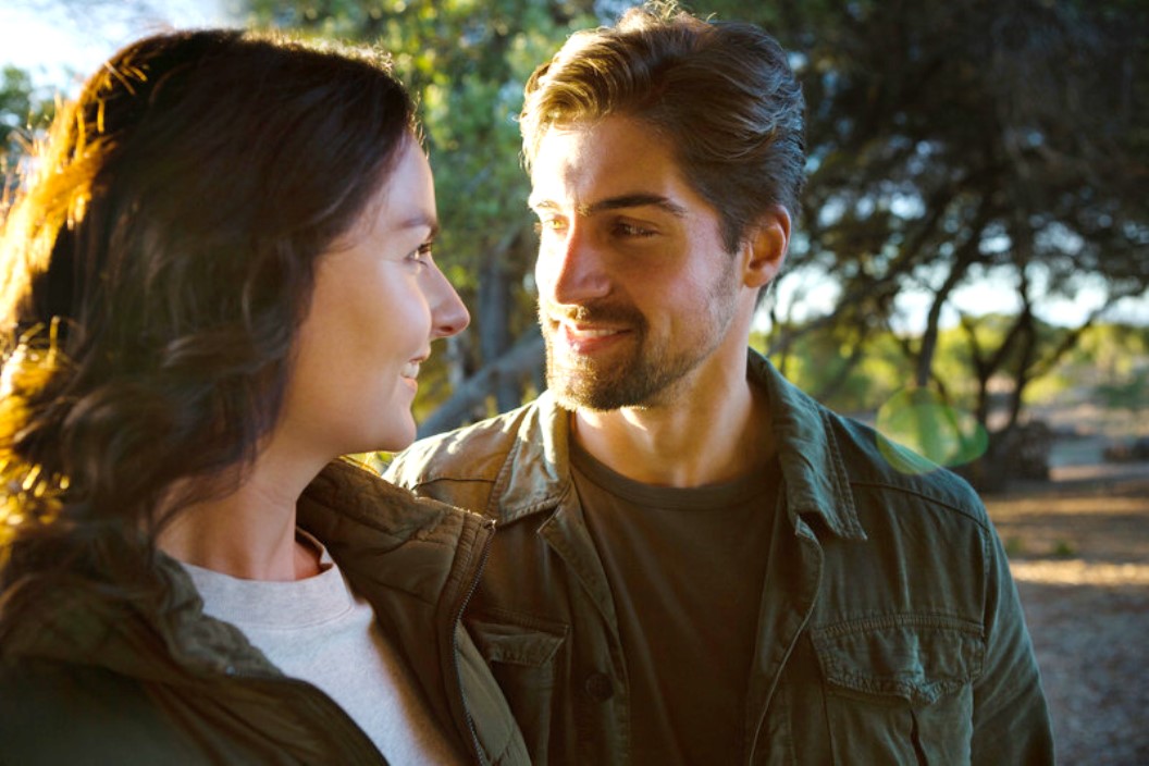 A man with short brown hair and a beard smiles at a woman with long brown hair in an outdoor setting. Both are dressed casually in green jackets, standing close together with trees and sunlight filtering through the background, creating a serene atmosphere.